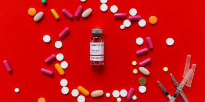 Colorful tablets and COVID-19 vaccine vial on a red background with syringes, symbolizing healthcare.